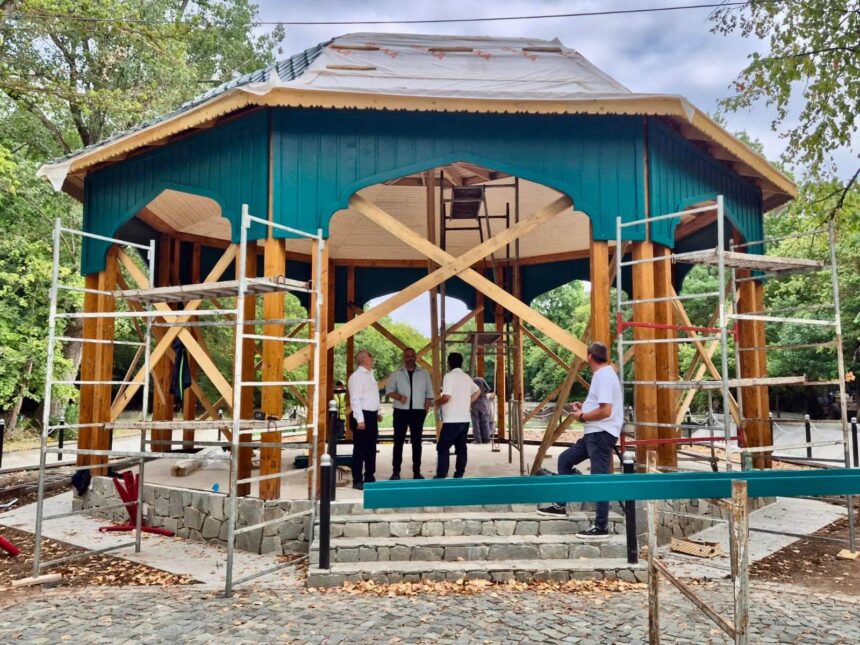 Under-construction wooden pavilion with teal siding in a park; scaffolding surrounds the structure and several workers stand nearby.