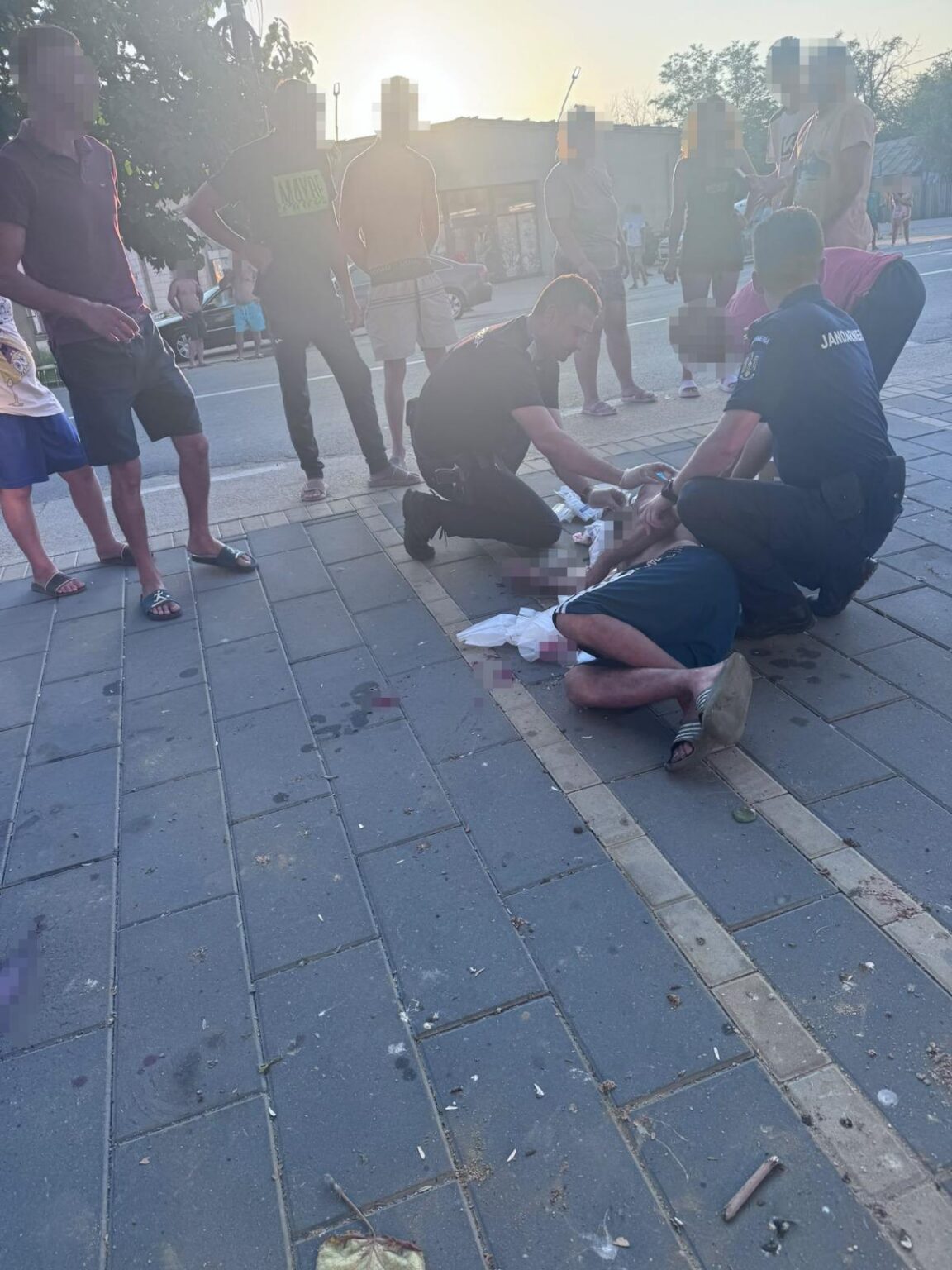 scarisoara WhatsApp Image 2026-08-11 at 101753s - Jurnalul Olteniei – Știri din Craiova și Oltenia Two officers kneel beside a man lying injured on a sidewalk while bystanders watch on a sunny urban street.