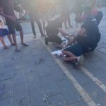 Two officers kneel beside a man lying injured on a sidewalk while bystanders watch on a sunny urban street.