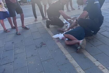 Two officers kneel beside a man lying injured on a sidewalk while bystanders watch on a sunny urban street.