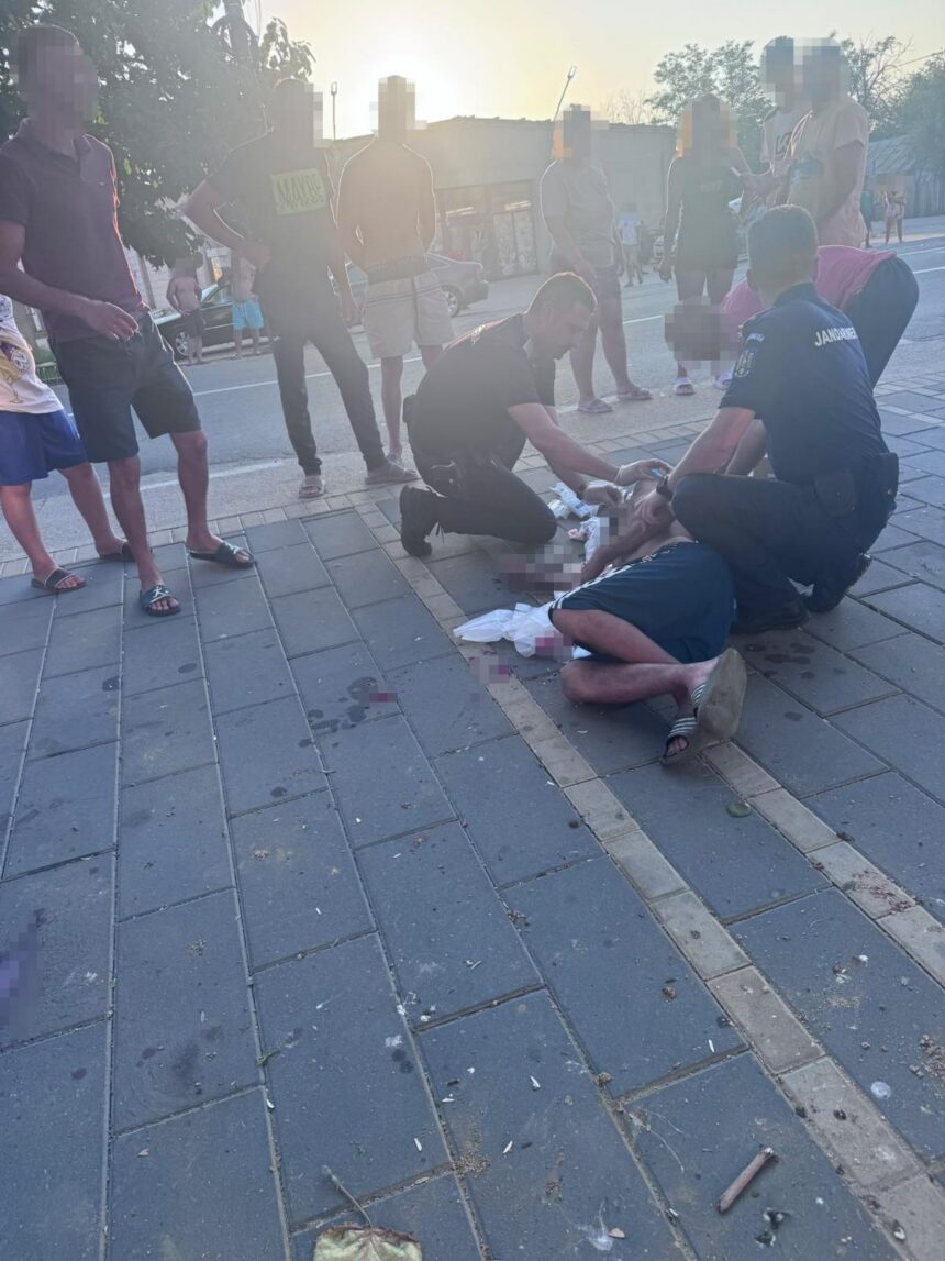 Two officers kneel beside a man lying injured on a sidewalk while bystanders watch on a sunny urban street.