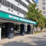 Hospital entrance with teal sign reading 'Spitalul Judetean de Urgenta', glass doors and a shaded drop-off area.