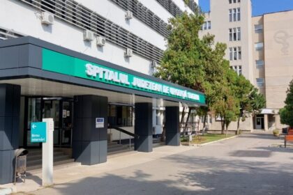 Hospital entrance with teal sign reading 'Spitalul Judetean de Urgenta', glass doors and a shaded drop-off area.