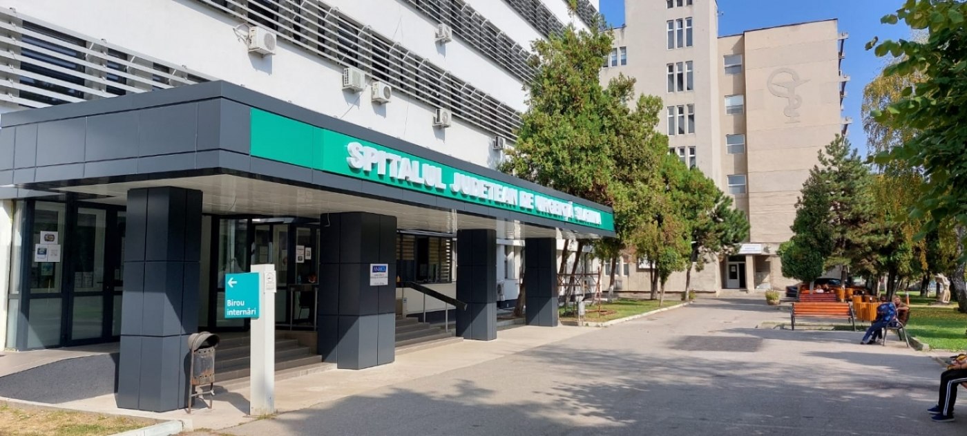 spitalul slatina index - Jurnalul Olteniei – Știri din Craiova și Oltenia Hospital entrance with teal sign reading 'Spitalul Judetean de Urgenta', glass doors and a shaded drop-off area.