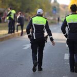 Two police officers in high-visibility jackets walk away down a city street as bystanders stand behind a cordoned area in the background.