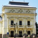 Ornate cream theater building facade with sculpted figures and decorative carvings under a blue sky, sign reads TEATR NATIONAL (theatre).