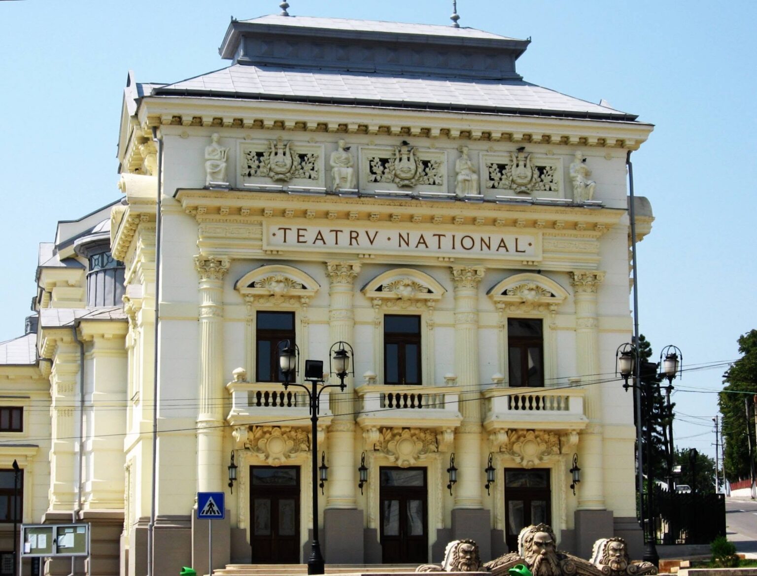 Ornate cream theater building facade with sculpted figures and decorative carvings under a blue sky, sign reads TEATR NATIONAL (theatre).