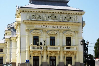 Ornate cream theater building facade with sculpted figures and decorative carvings under a blue sky, sign reads TEATR NATIONAL (theatre).