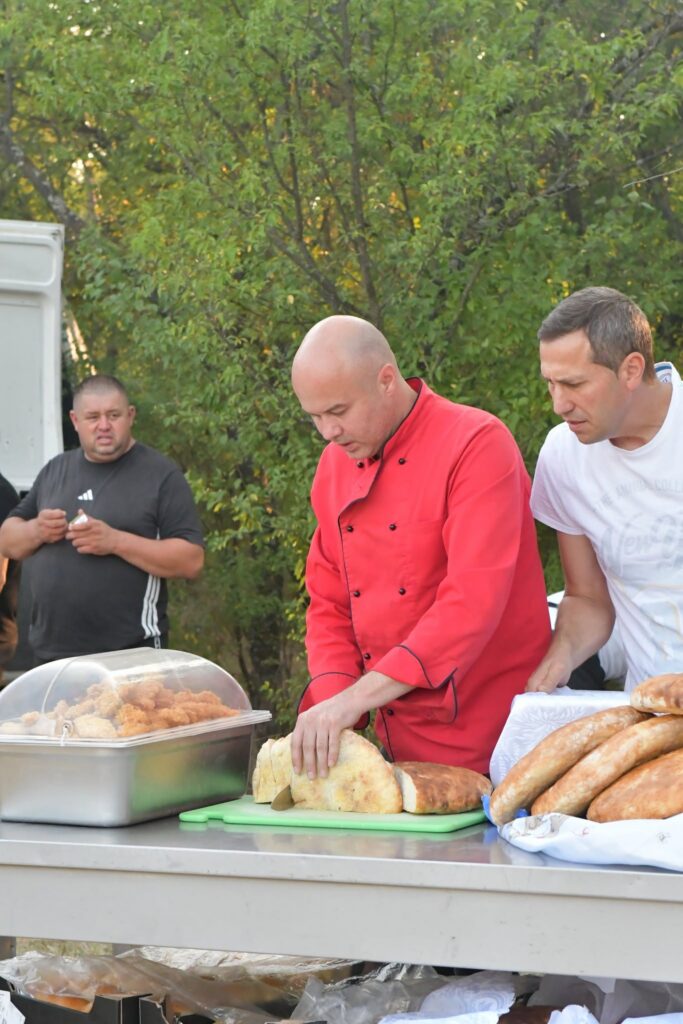 Chef in a red uniform serves bread rolls at an outdoor food stall, with a tray of fried foods and buns on the stainless table.