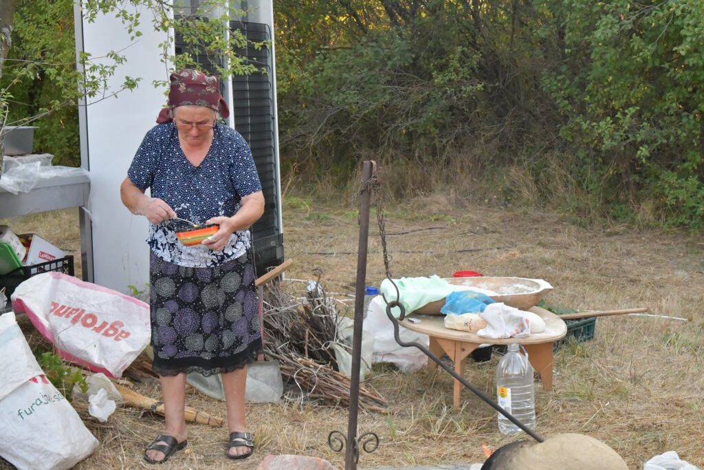 Elderly woman wearing a burgundy headscarf and glasses, mixing food in a striped bowl outdoors beside a caravan and scattered supplies.