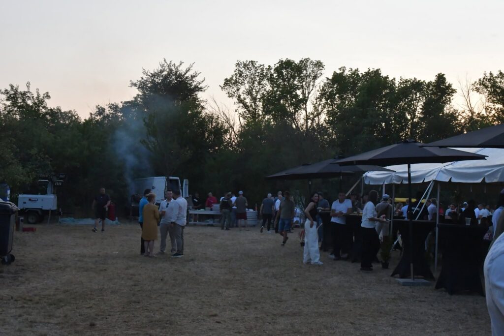 Outdoor social gathering in a field with people, tents, and tables at dusk; trees in the background.