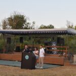 Outdoor stage setup with a blue-and-white canopy, speakers, and performers at a microphone and podium, with trees in the background.