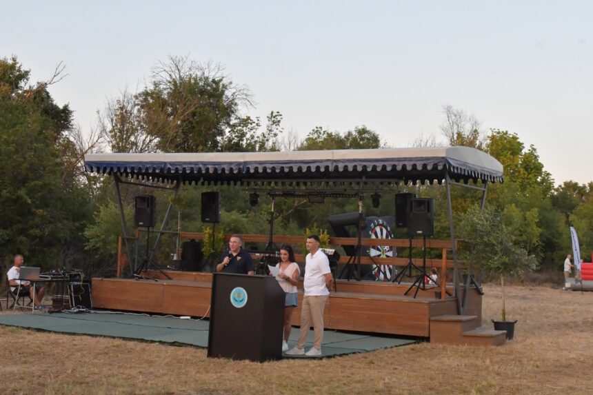 Outdoor stage setup with a blue-and-white canopy, speakers, and performers at a microphone and podium, with trees in the background.