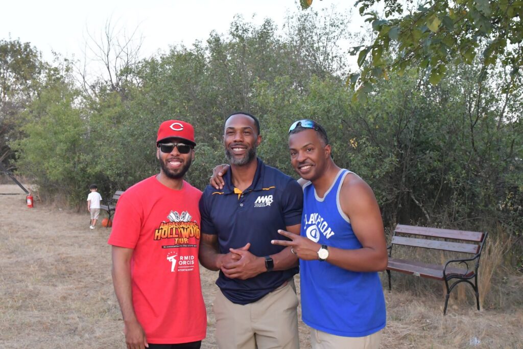 Three men posing together outdoors in a park, smiling at the camera, dressed in red, navy, and blue athletic shirts with trees in the background.