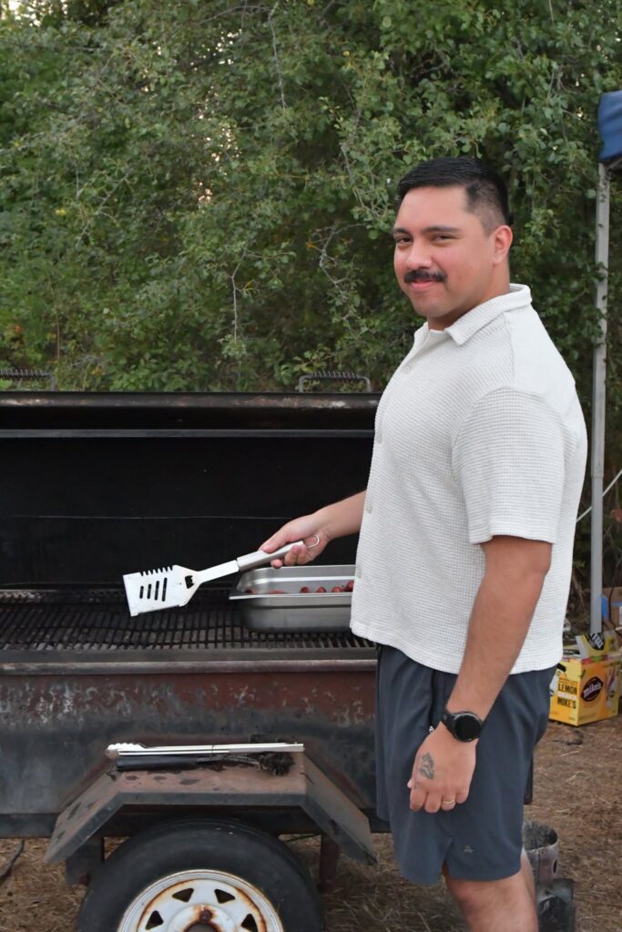 Man in a light polo shirt grilling outdoors, holding a spatula over a barbecue grill with food nearby.