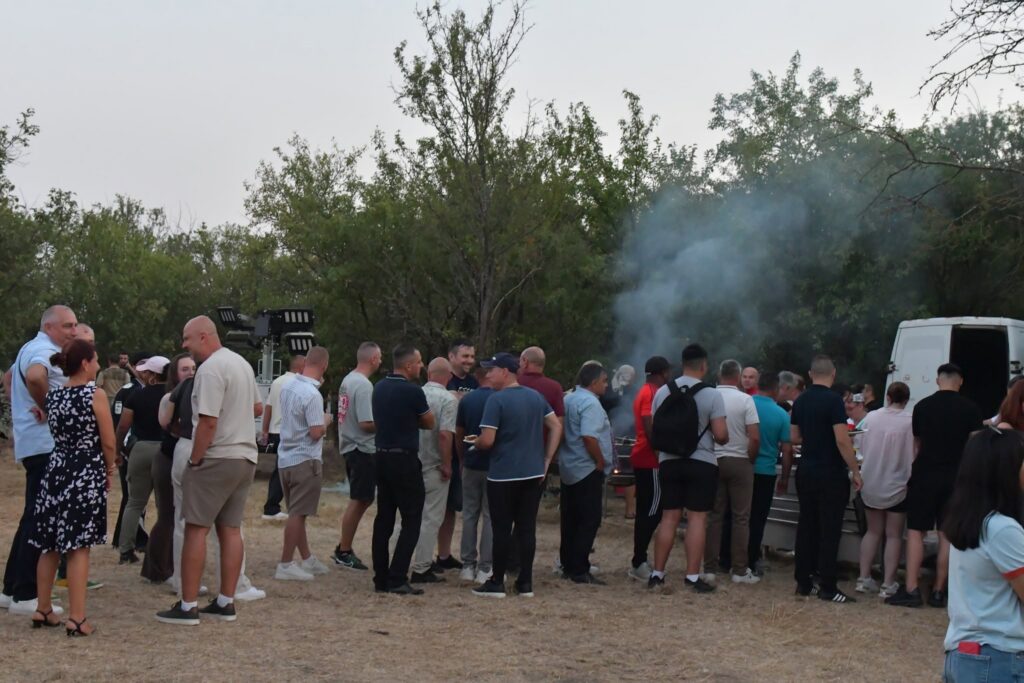 Crowd of people outdoors at a gathering, some talking, with smoke from a barbecue and a white van nearby in the background.