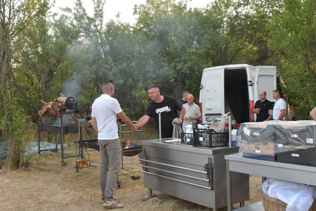 Group of men cooking barbecue outdoors with a metal rotisserie grill and smoked meat, near a white van.