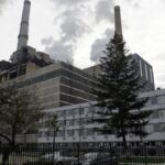 Industrial power plant with tall smokestacks emitting smoke, adjacent gray multi-story building, trees, and a fence in the foreground