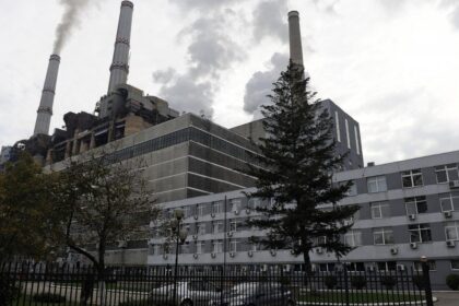 Industrial power plant with tall smokestacks emitting smoke, adjacent gray multi-story building, trees, and a fence in the foreground