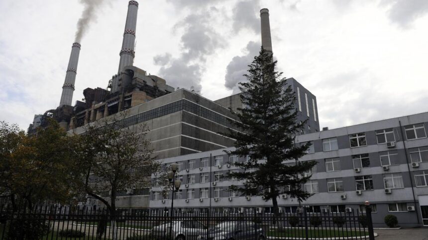 Industrial power plant with tall smokestacks emitting smoke, adjacent gray multi-story building, trees, and a fence in the foreground