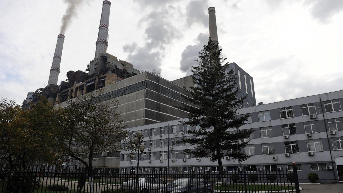 Industrial power plant with tall smokestacks emitting smoke, adjacent gray multi-story building, trees, and a fence in the foreground