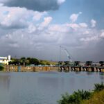 Industrial waterfront scene with a tilted blue crane over a river, low buildings on the left, and cloudy sky above.