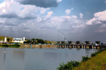 Industrial waterfront scene with a tilted blue crane over a river, low buildings on the left, and cloudy sky above.