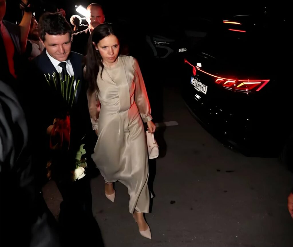 A man in a dark suit and tie carrying flowers walks beside a woman in a beige dress at night, while photographers flash from behind and a dark car with red taillights is nearby.