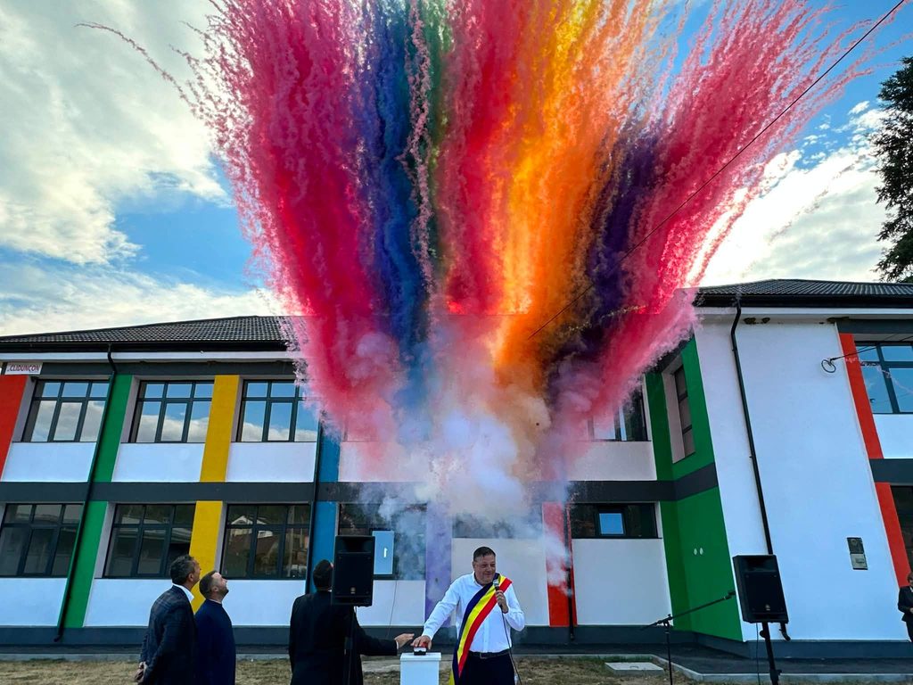 Rainbow-colored smoke bursts over a modern building during an outdoor ceremony, with onlookers and a man wearing a ceremonial sash nearby.
