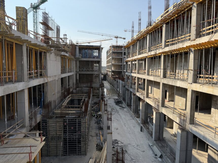 Large construction site with concrete structures, scaffolding, and tower cranes under a clear blue sky