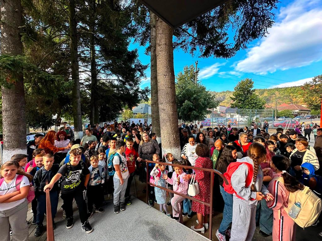Large crowd of schoolchildren waiting in a line behind a railing outdoors, with trees and a blue sky in the background.
