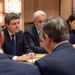 Group of professionals in a meeting around a conference table; man in a blue suit speaks as others listen.