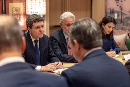 Group of professionals in a meeting around a conference table; man in a blue suit speaks as others listen.