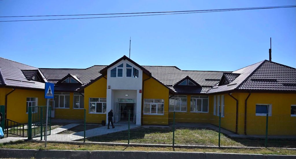 799725922_1120225140487425_6768775401015272705_n - Jurnalul Olteniei – Știri din Craiova și Oltenia Bright yellow school-like building with a white central entrance and peaked roof, under a blue sky, fence in front.