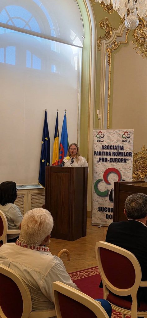 Female presenter at a wooden podium delivering a speech in an ornate room with flags behind her and a banner reading 'Asociatia Partida Romilor Pro-Europa'.