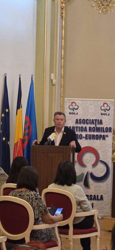 Man in a suit giving a speech at a wooden podium, with EU and Romanian flags and a colorful banner behind him, in a formal room with seated audience in front.