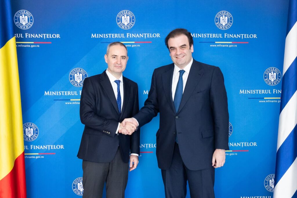 Two men in dark suits shake hands in front of a blue backdrop with government seals and Romanian flags in the corners, during a formal event at the Ministry of Finance.