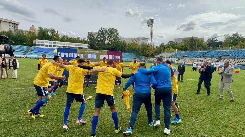 Soccer players in yellow jerseys form a huddle on a grass field after a match, with blue-clad staff nearby.