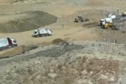 Construction site with dirt piles and several trucks, including white dump trucks, on a dusty excavated area.