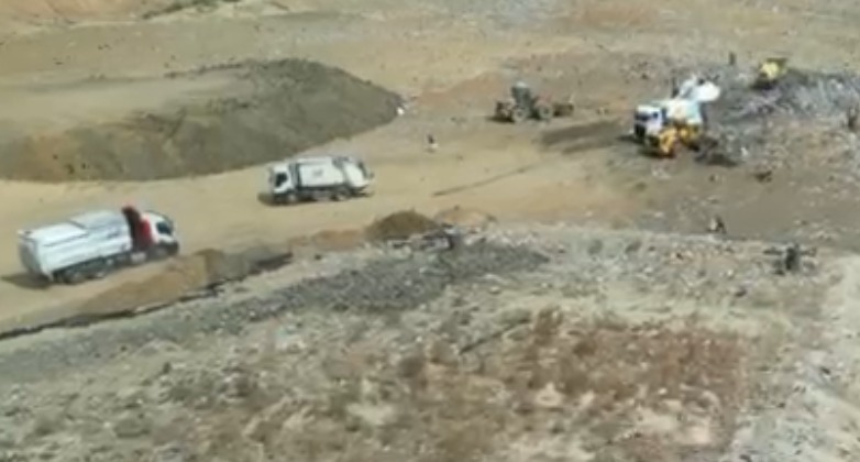 Construction site with dirt piles and several trucks, including white dump trucks, on a dusty excavated area.