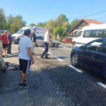 Group of bystanders and responders near a car crash on a sunny street; an ambulance and a damaged car are present.