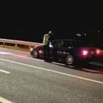 Nighttime traffic stop: a police officer in a reflective vest speaks to the driver of a dark sedan on a highway shoulder near a guardrail.