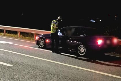 Nighttime traffic stop: a police officer in a reflective vest speaks to the driver of a dark sedan on a highway shoulder near a guardrail.