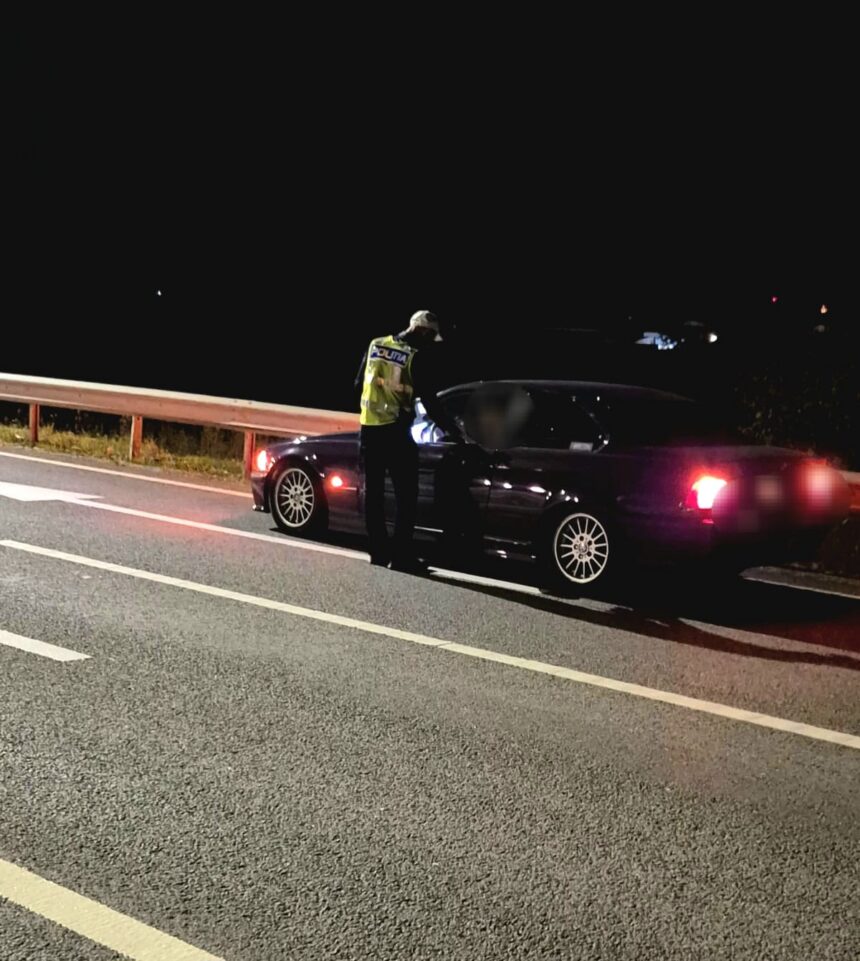 Nighttime traffic stop: a police officer in a reflective vest speaks to the driver of a dark sedan on a highway shoulder near a guardrail.