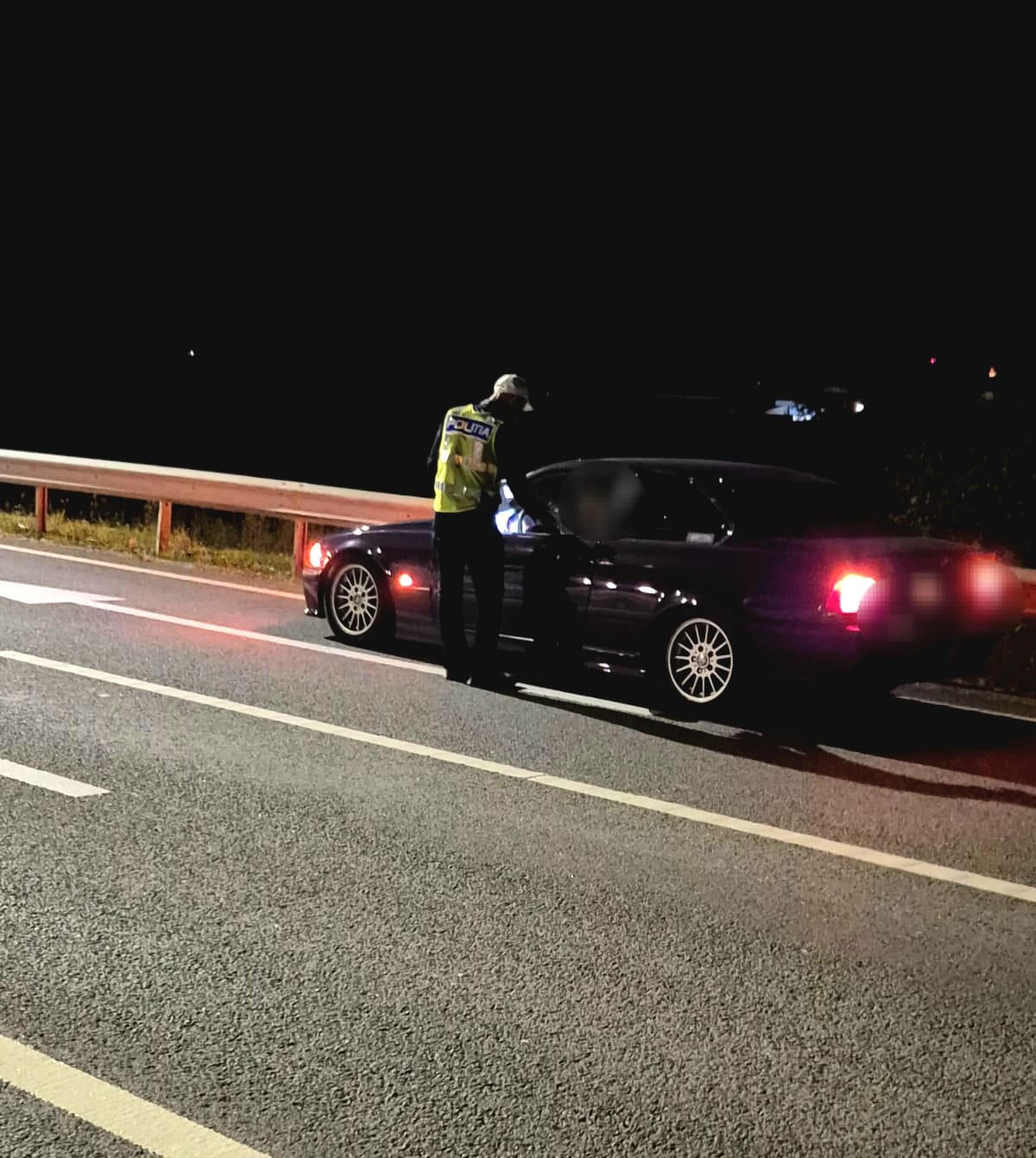 Nighttime traffic stop: a police officer in a reflective vest speaks to the driver of a dark sedan on a highway shoulder near a guardrail.