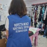 Back view of a person wearing a blue vest with white text, likely an inspector, standing in a room with clothing racks visible in the background.