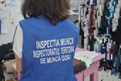 Back view of a person wearing a blue vest with white text, likely an inspector, standing in a room with clothing racks visible in the background.