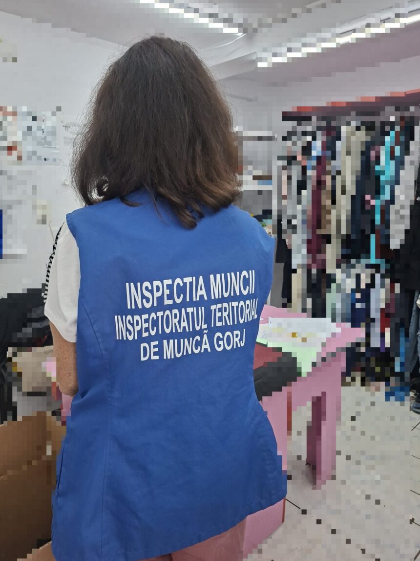 Back view of a person wearing a blue vest with white text, likely an inspector, standing in a room with clothing racks visible in the background.