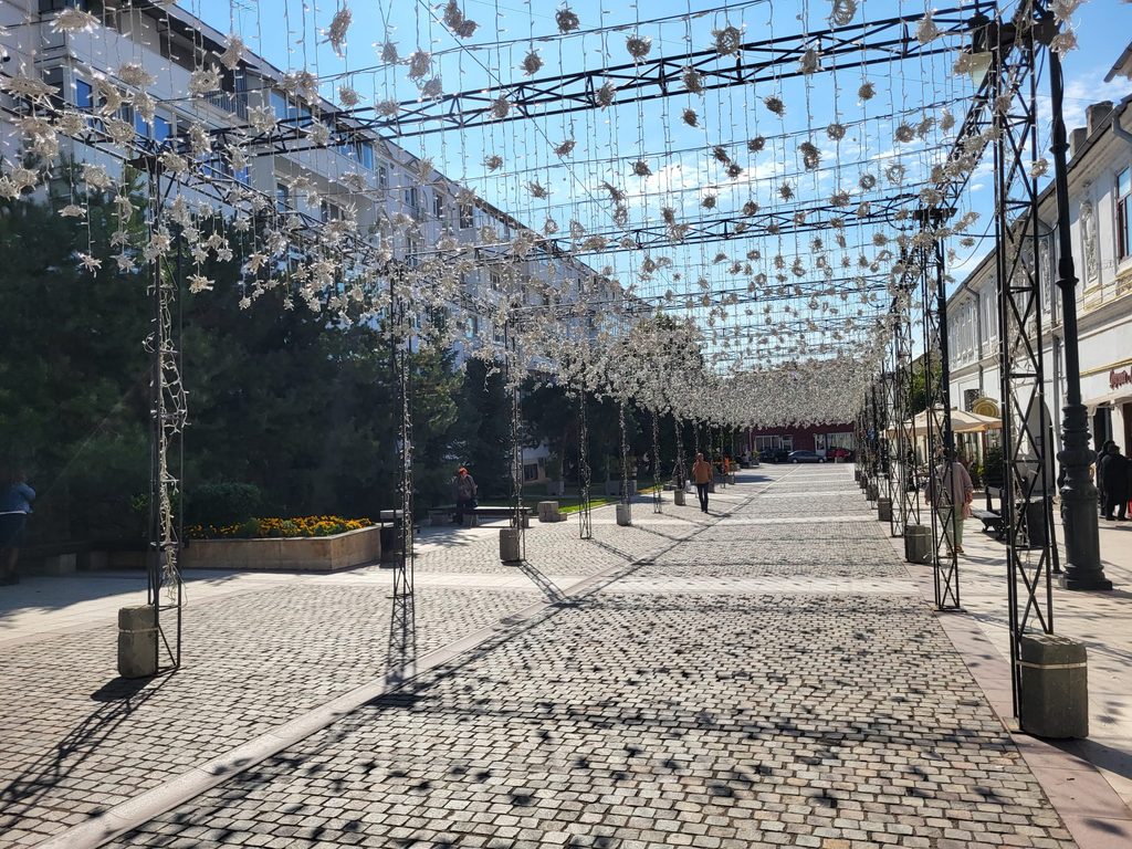 Outdoor pedestrian street under a decorative white light canopy of strings and flowers over a cobblestone path with people walking nearby.