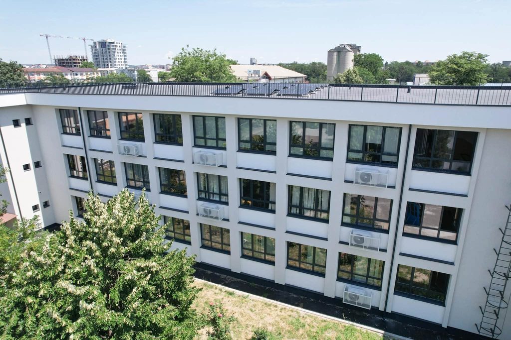 High-angle view of a modern white apartment building with large dark-framed windows and solar panels on the flat roof, with green trees in the foreground.